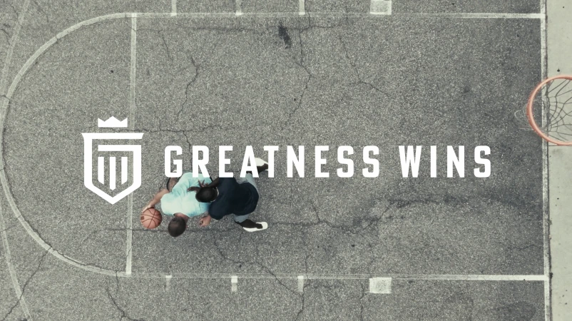 Overhead view of two adults playing basketball on an outdoor court with the text "Greatness Wins" and a white logo.