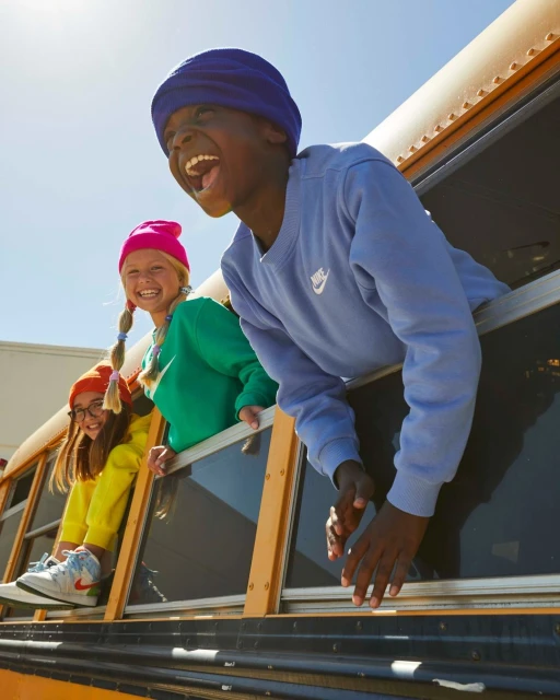 Three happy children lean out of a yellow school bus window, wearing bright colored clothes and hats, and show a lot of teeth as they laugh.