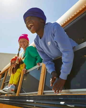 Three happy children lean out of a yellow school bus window, wearing bright colored clothes and hats, and show a lot of teeth as they laugh.