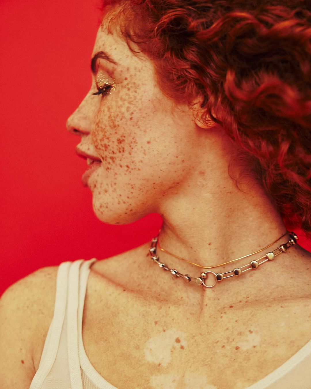 Profile portrait of a red-haired woman with freckles on a red background. She wears gold glitter eye makeup and delicate necklaces.