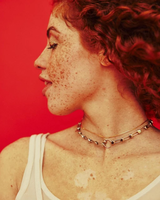 Profile portrait of a red-haired woman with freckles on a red background. She wears gold glitter eye makeup and delicate necklaces.