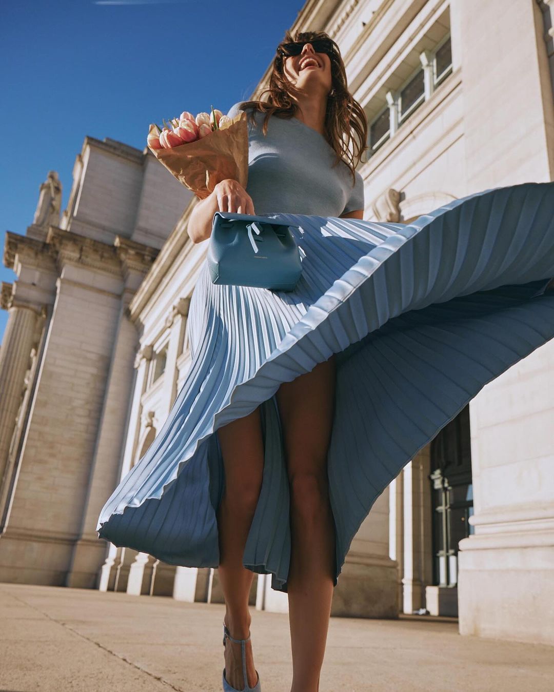 Smiling woman in a light blue pleated skirt walks on the sidewalk holding a bouquet of flowers and a light blue purse.