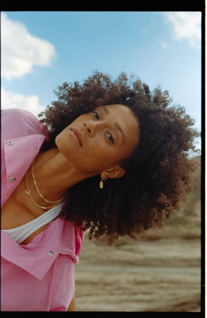 Portrait of an adult woman with curly dark hair and freckles, wearing gold jewelry and a pink shirt, posing in an outdoor setting.