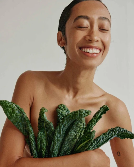 Adult woman with a beaming smile, holding dark green kale leaves covering her chest, against a neutral background.