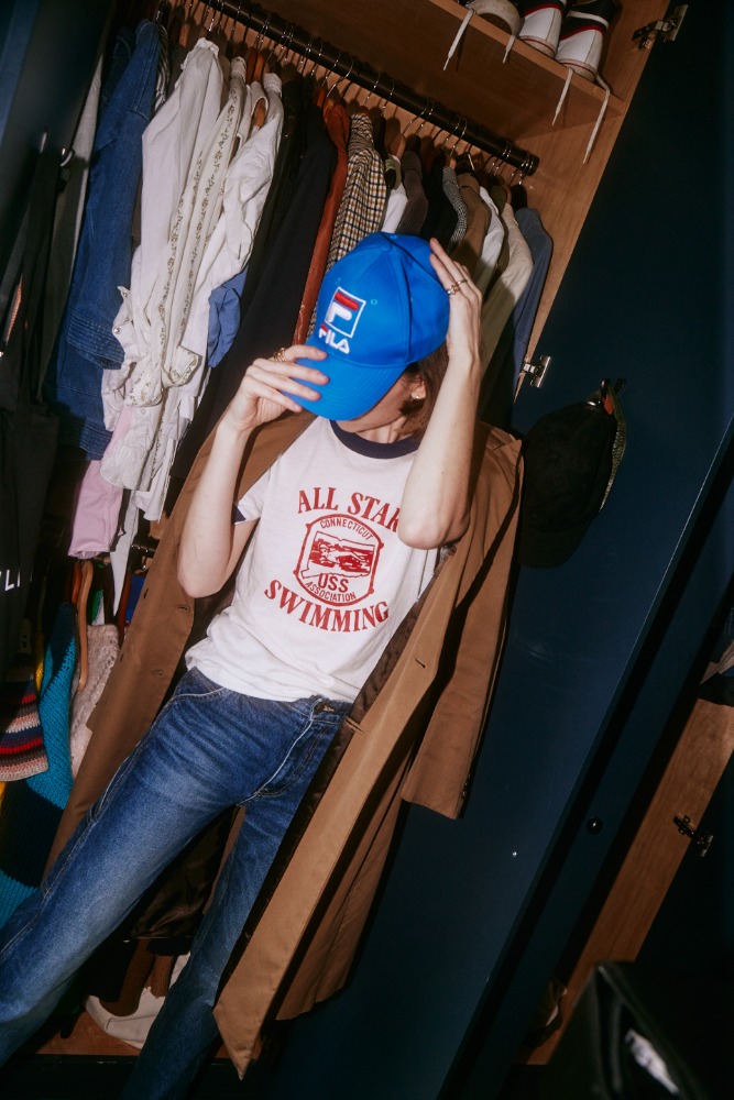 Person in blue Fila cap, white t-shirt, and blue jeans stands in front of a wardrobe with clothes on hangers and on top shelf.