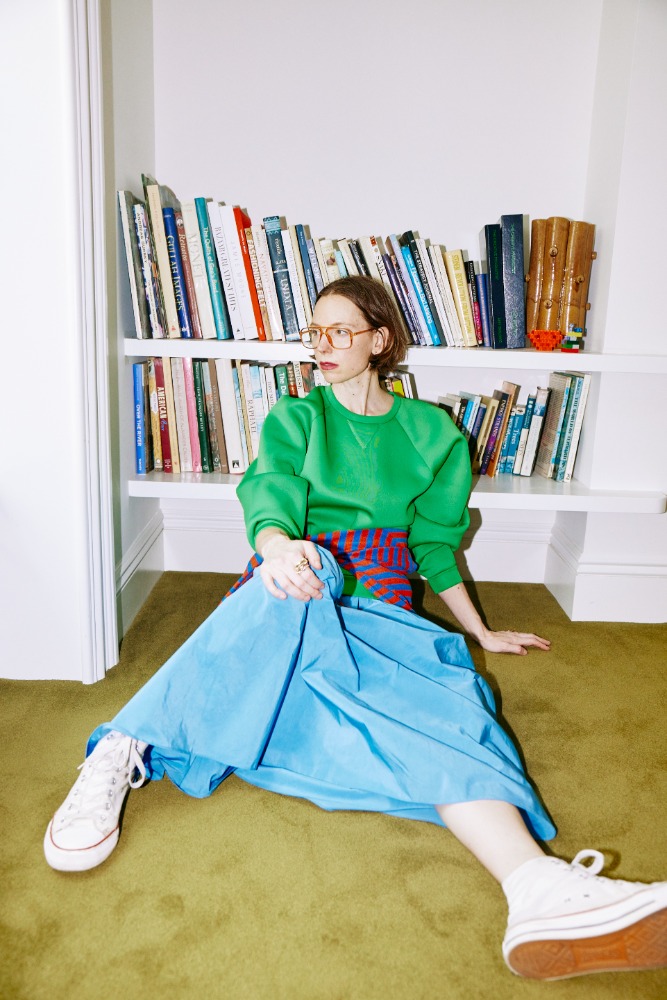 A woman sits on a carpeted floor in front of a bookshelf, wearing a green sweatshirt, blue skirt, and white canvas sneakers.