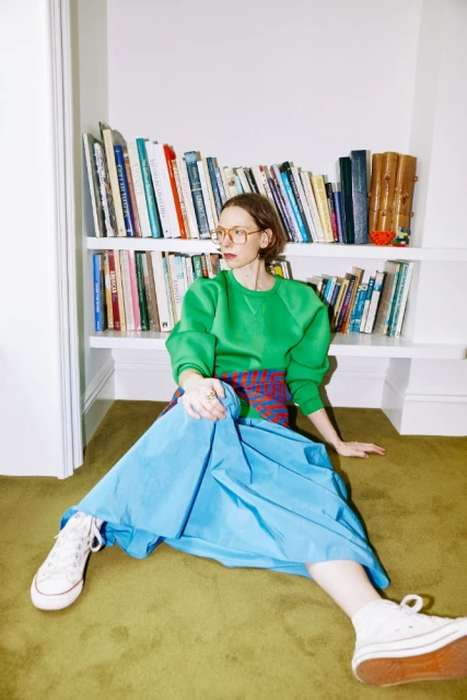 A woman sits on a carpeted floor in front of a bookshelf, wearing a green sweatshirt, blue skirt, and white canvas sneakers.