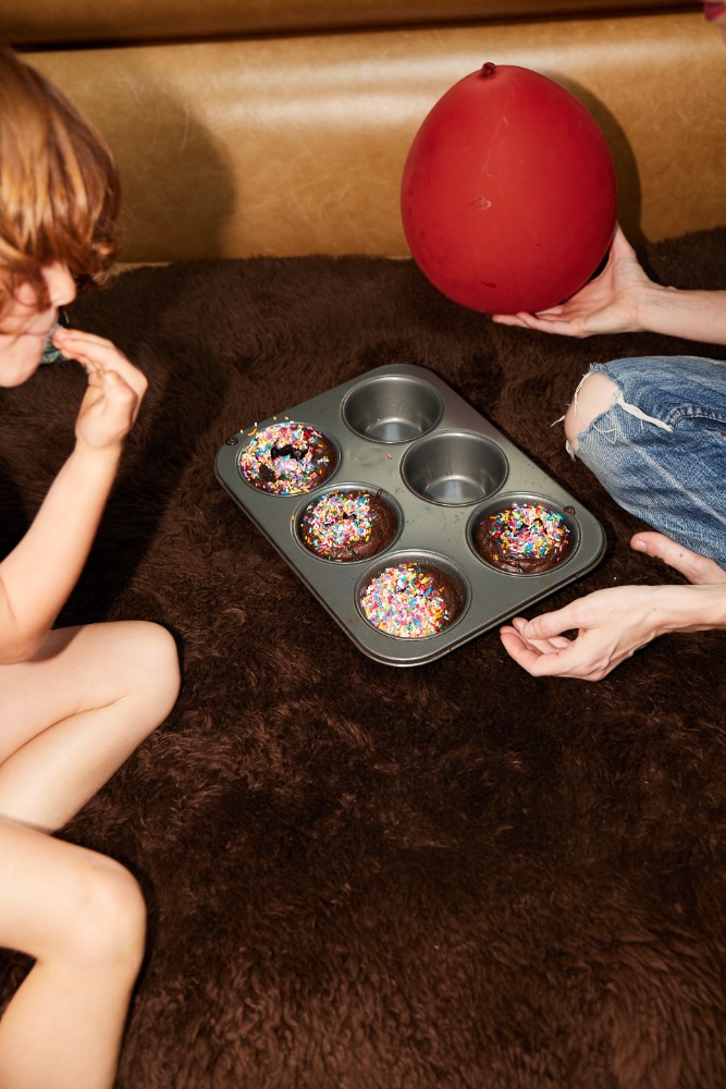 Two people near a muffin tin of decorated chocolate cupcakes and a red balloon on a textured brown surface indoors.