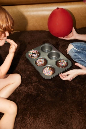 Two people near a muffin tin of decorated chocolate cupcakes and a red balloon on a textured brown surface indoors.