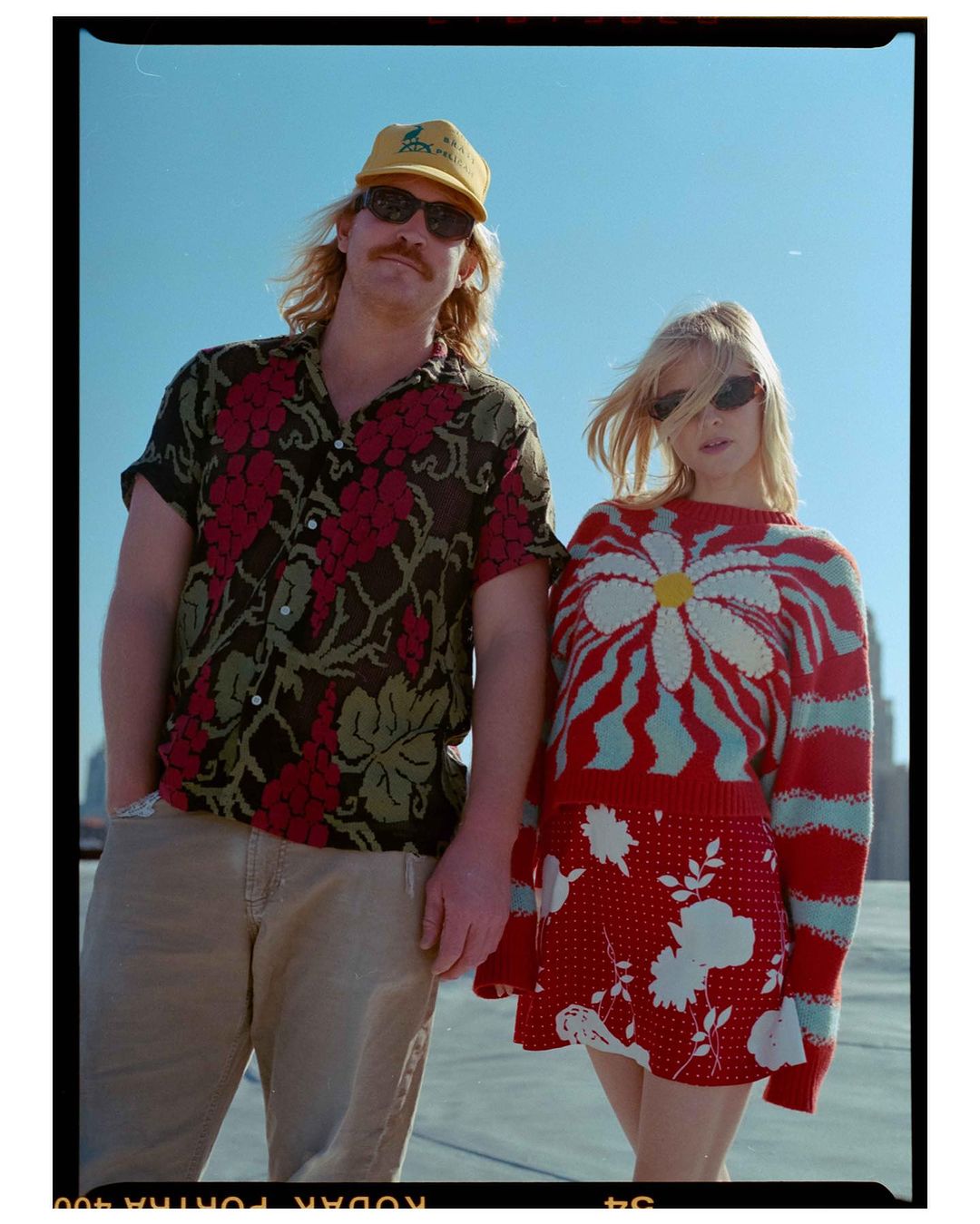 A stylish couple poses on a rooftop in bright sunlight, the woman in a floral mini-skirt and daisy sweater, the man in a floral shirt and cap.