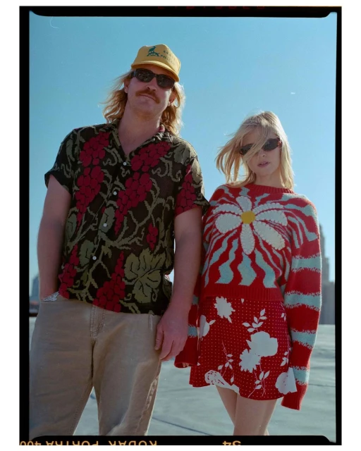 A stylish couple poses on a rooftop in bright sunlight, the woman in a floral mini-skirt and daisy sweater, the man in a floral shirt and cap.