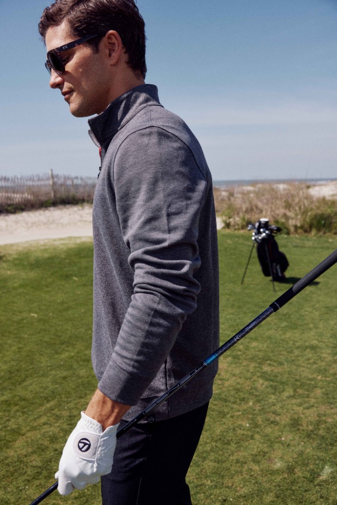 An adult male golfer on a sunny day, wearing sunglasses and a grey sweater, holding a golf club, with the ocean in the background.