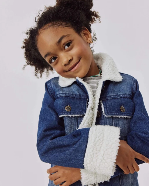 Portrait of a smiling child in a denim jacket with a sherpa collar against a neutral background in a studio setting. 