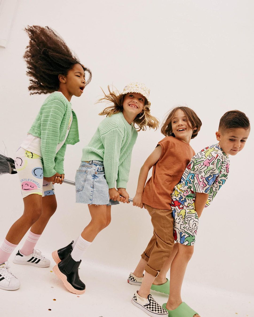 Four smiling children in fashionable casual clothing 'fly' on a broomstick in a light studio against a white backdrop; playful, fun.