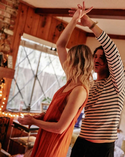 A couple dancing indoors with arms raised; the woman wears an orange dress while the man wears a striped shirt with a mustache.