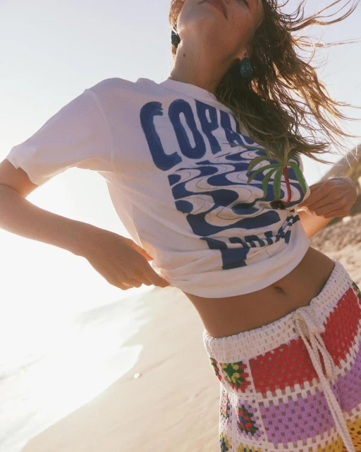 A woman stands on a beach in bright sunlight, wearing a graphic tee and crochet shorts, with blue earrings and wind-blown hair.