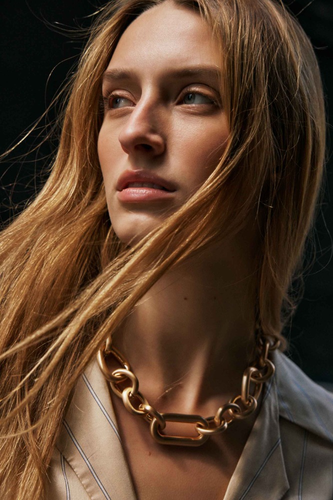 Close up portrait of a blonde woman wearing a chunky gold chain necklace and a striped beige shirt against a dark background