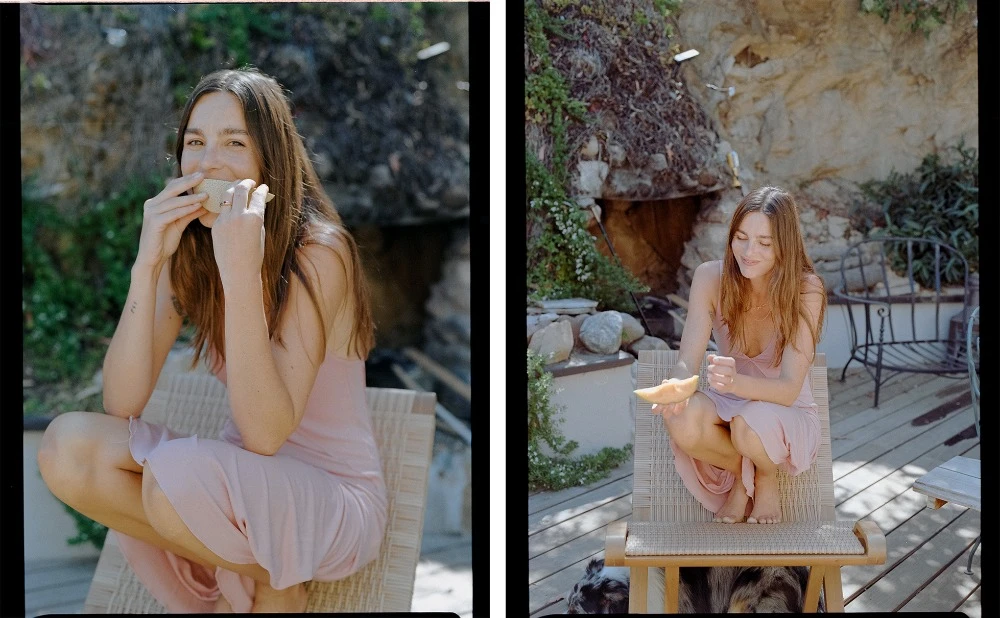 A woman in a pink dress is sitting on a wicker chair eating a wrap outside, while another woman eats a slice of melon.