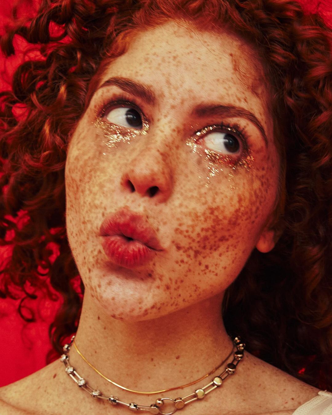 Close-up portrait of a woman with red curly hair, heavy freckles, and glitter makeup around her eyes against a red background.