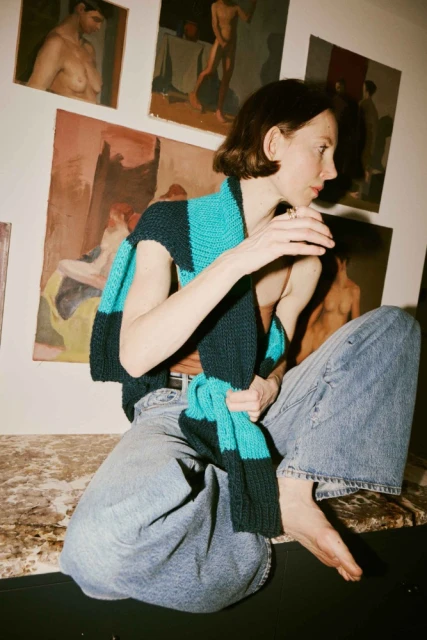 A woman with short brown hair sits on a black surface indoors wearing a striped blue and black scarf and blue jeans, with paintings in the background.