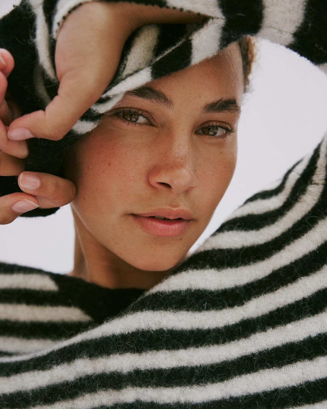 Close-up portrait of a woman wearing a black and white striped sweater with hands framing her face against a white background.