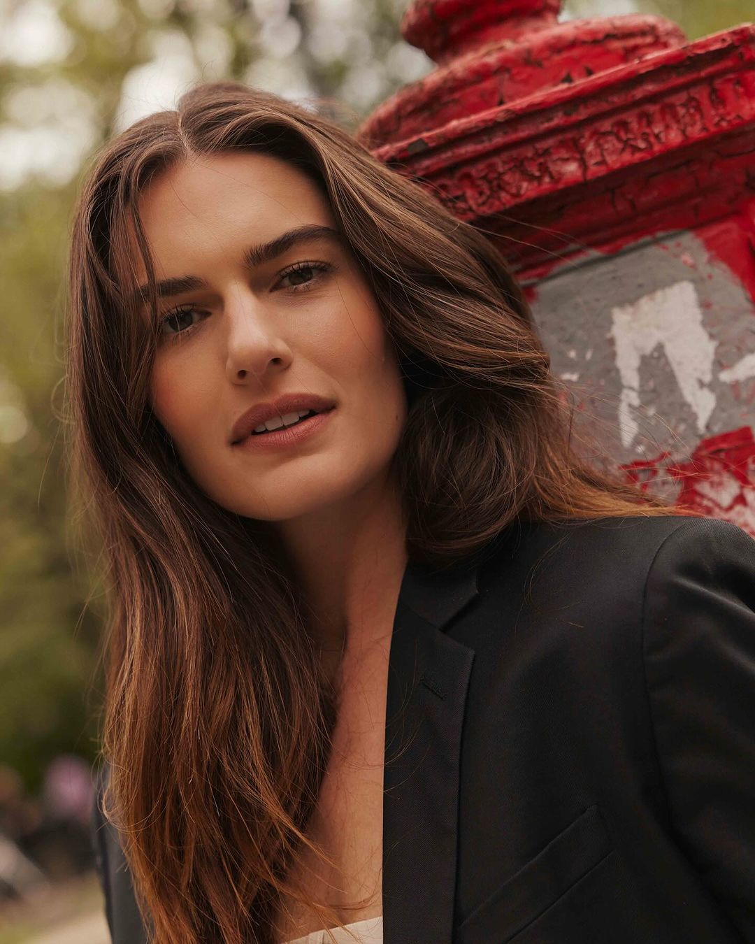 Portrait of a brunette woman in a black blazer leaning against a red fire hydrant, with soft natural lighting and a shallow depth of field.