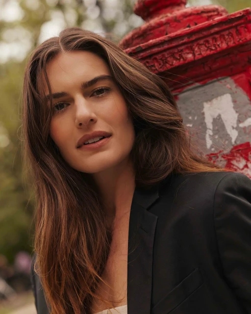 Portrait of a brunette woman in a black blazer leaning against a red fire hydrant, with soft natural lighting and a shallow depth of field.