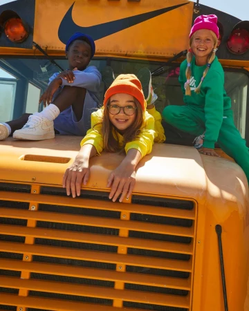 Three children pose atop a yellow school bus, wearing bright, colorful outfits and matching knit hats, with the Nike logo visible above.