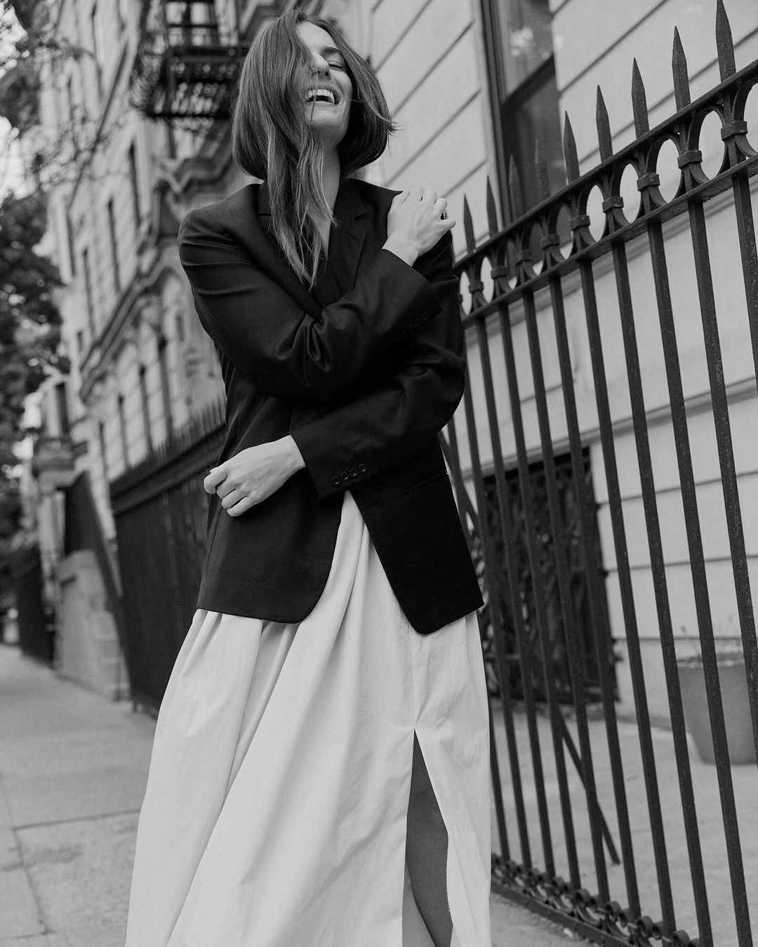 A woman stands on a city sidewalk in a black blazer and a tan maxi skirt. She has a cheerful facial expression.