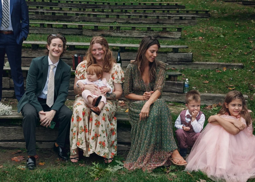 A group of casually dressed adults and children sit on bleachers covered in grass in a grassy outdoor setting, enjoying a relaxed gathering.
