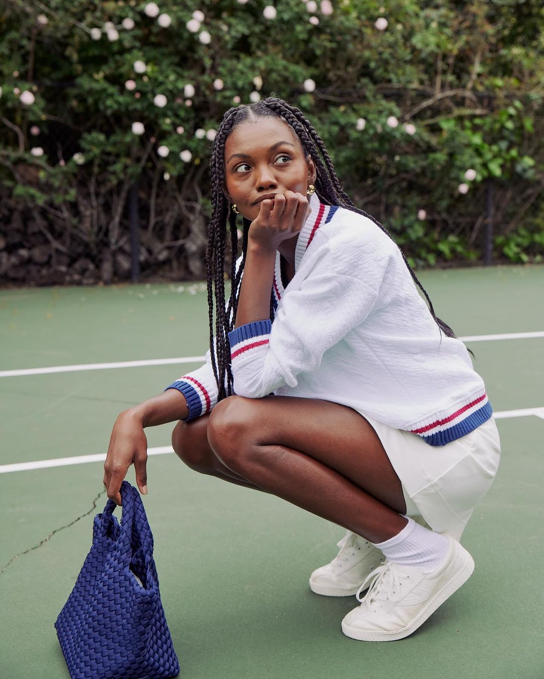 A dark-skinned woman with braids wearing athletic wear, crouching on a tennis court, holding a woven blue bag, outdoors on a sunny day.