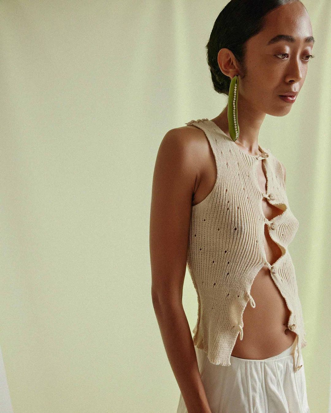 Young woman in a cream, partially unbuttoned knit top and white pants poses against a pale green backdrop, accessorized with large earrings.