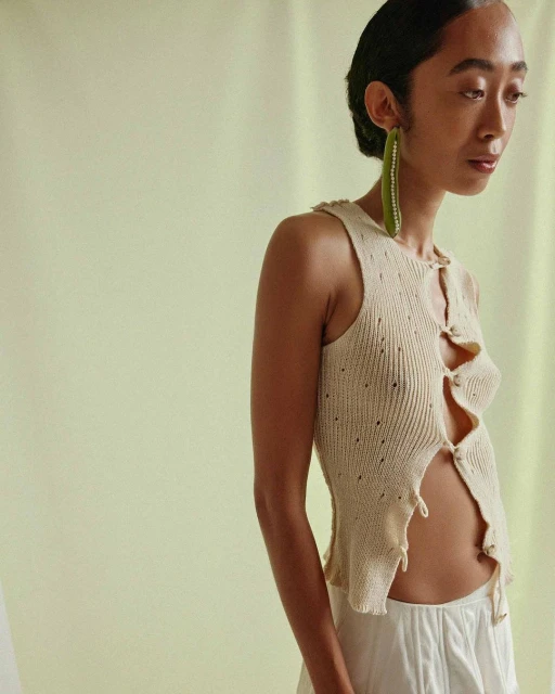Young woman in a cream, partially unbuttoned knit top and white pants poses against a pale green backdrop, accessorized with large earrings.
