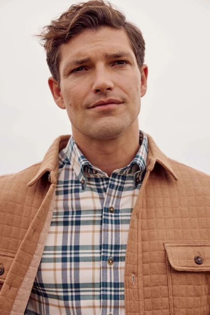 Portrait of a handsome man wearing a tan quilted jacket and blue and tan plaid shirt, standing outdoors with a neutral-tone sky.