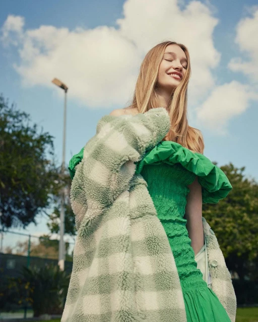 Blonde woman smiles in green dress and sherpa coat outdoors, posing against blue sky with clouds on sunny day.