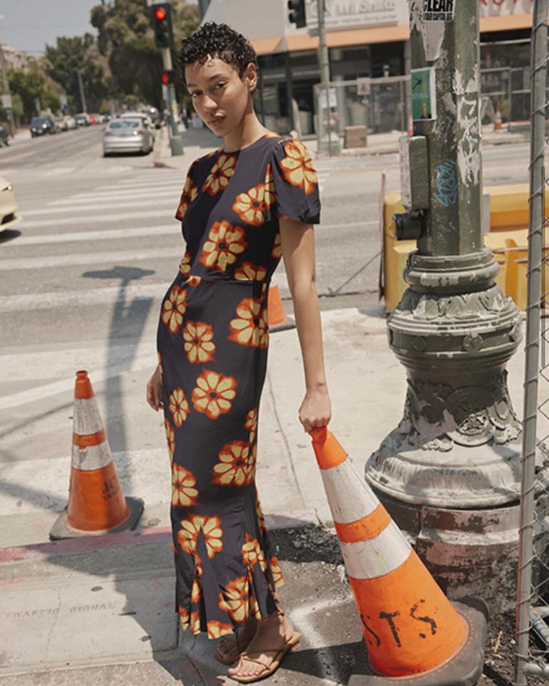 Woman in floral maxi dress stands on a city street holding a traffic cone, showcasing fashion apparel with urban environment in background.