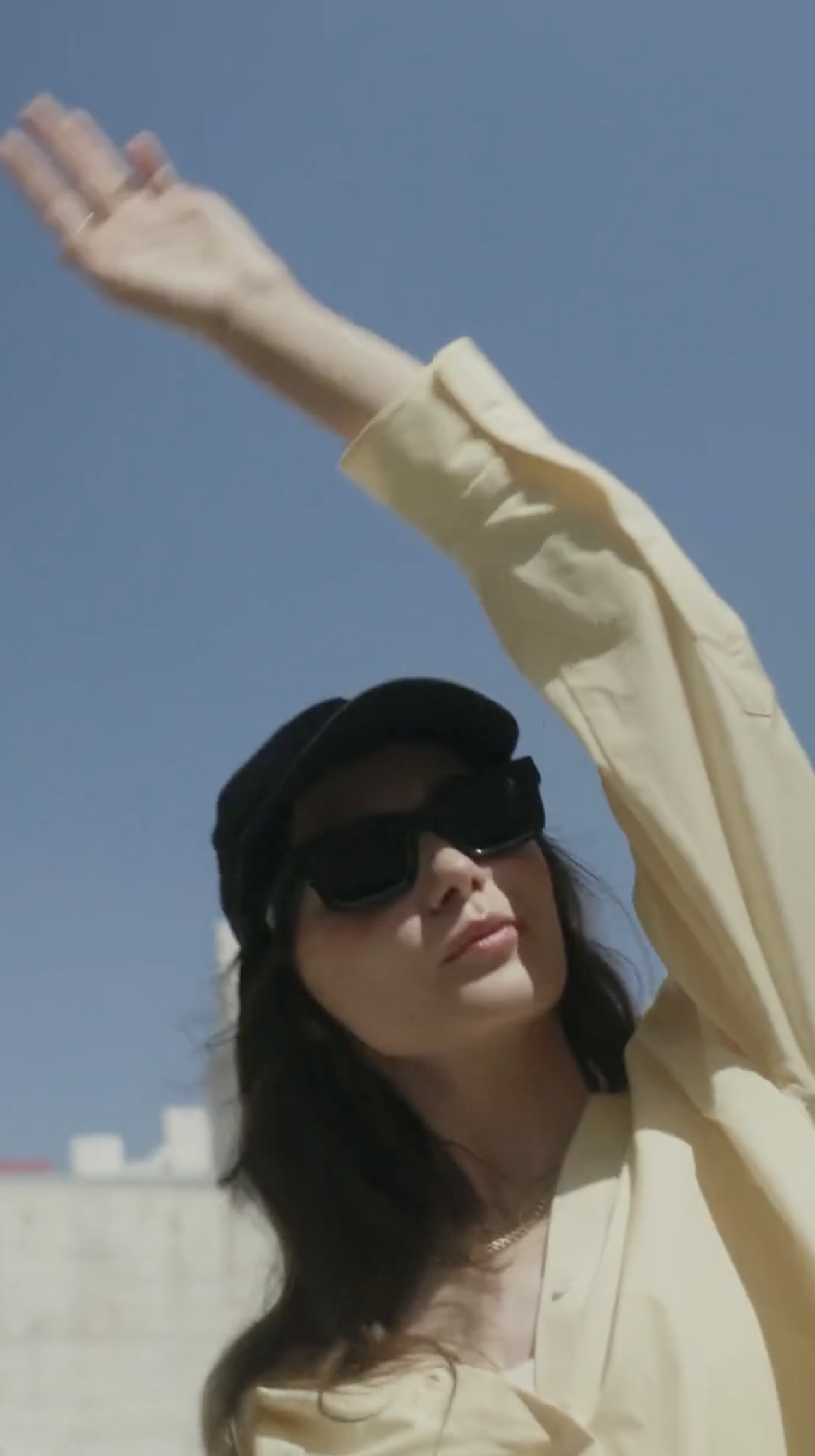 Woman in a yellow shirt and cap with raised arm against a clear blue sky, architectural structure in the background.