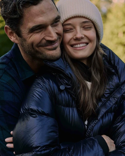 Smiling couple outdoors. A man with dark hair embraces a woman wearing a cream beanie and a navy puffer jacket in a nature setting.