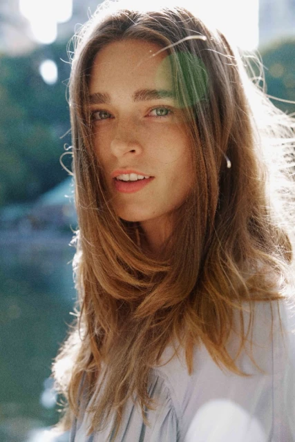 Close-up portrait of a young adult woman with long brown hair looking at the camera with a neutral expression in an outdoor setting.