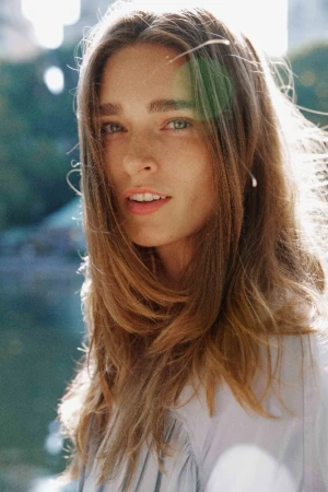 Close-up portrait of a young adult woman with long brown hair looking at the camera with a neutral expression in an outdoor setting.