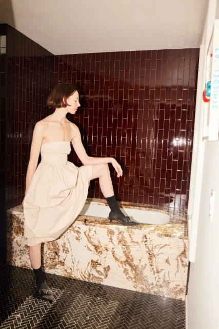 A fashionable young woman in a dress and socks poses in a bathroom with dark red brick walls and marble bathtub surround.