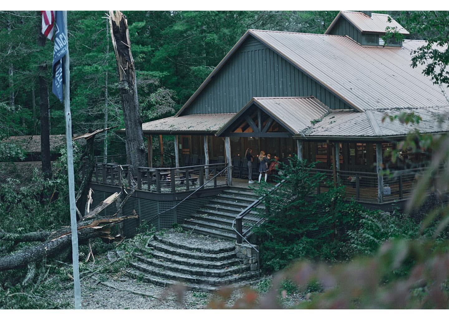Outdoor view of a group of people standing on the porch of a building with stairs, surrounded by lush green trees and a damaged tree.