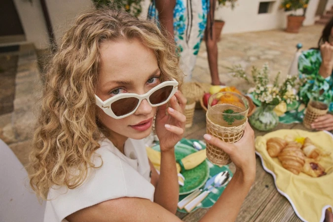 Close-up portrait of a woman with curly blonde hair wearing white sunglasses and holding a glass of iced drink at an outdoor table.