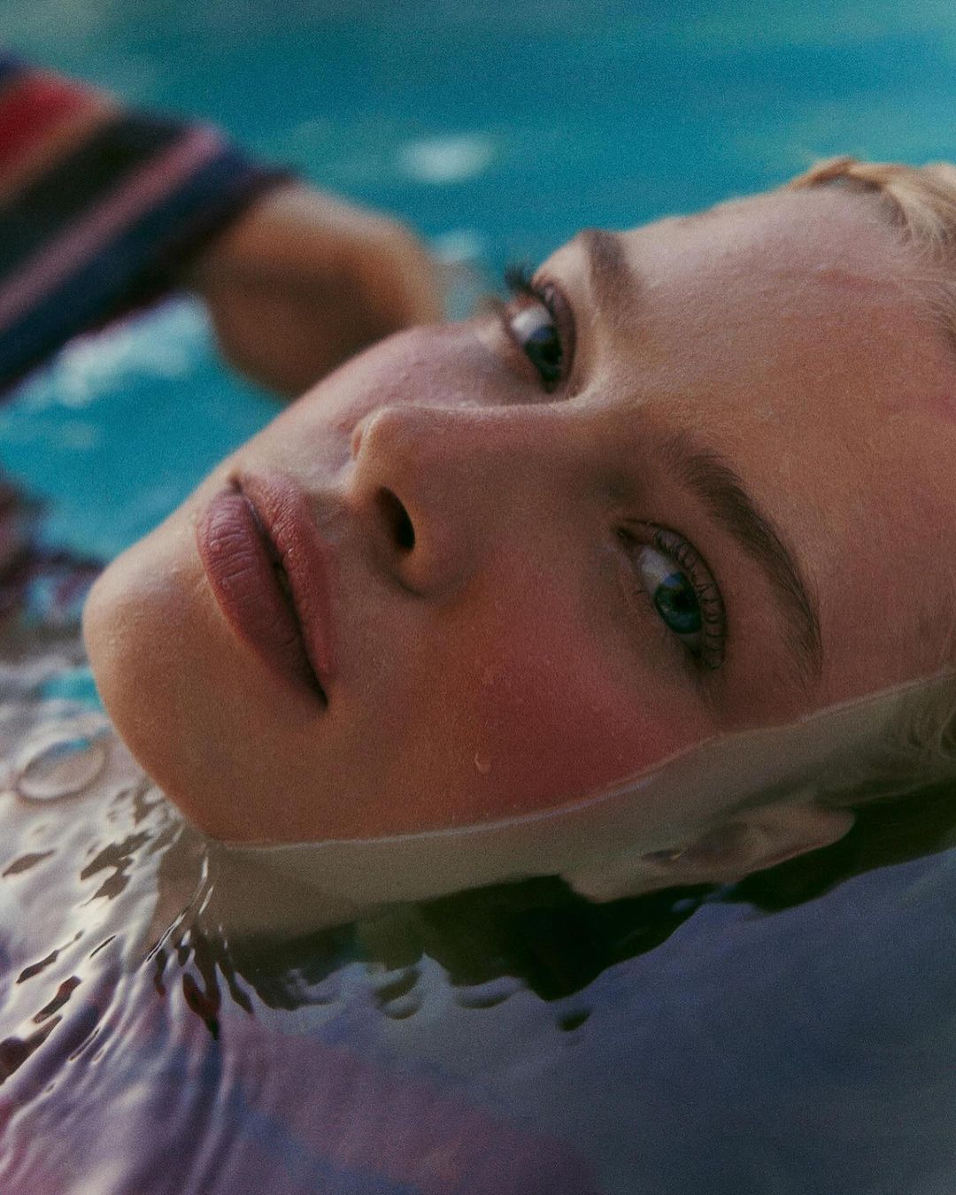 Close-up of a blond woman partially submerged in water, gazing upward with a serene expression, captured in soft, diffused light.
