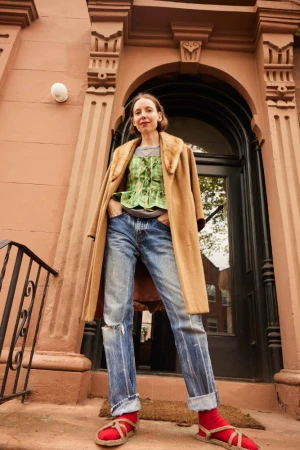 Woman in brown coat, green top, and distressed jeans standing before a brown architectural doorway.