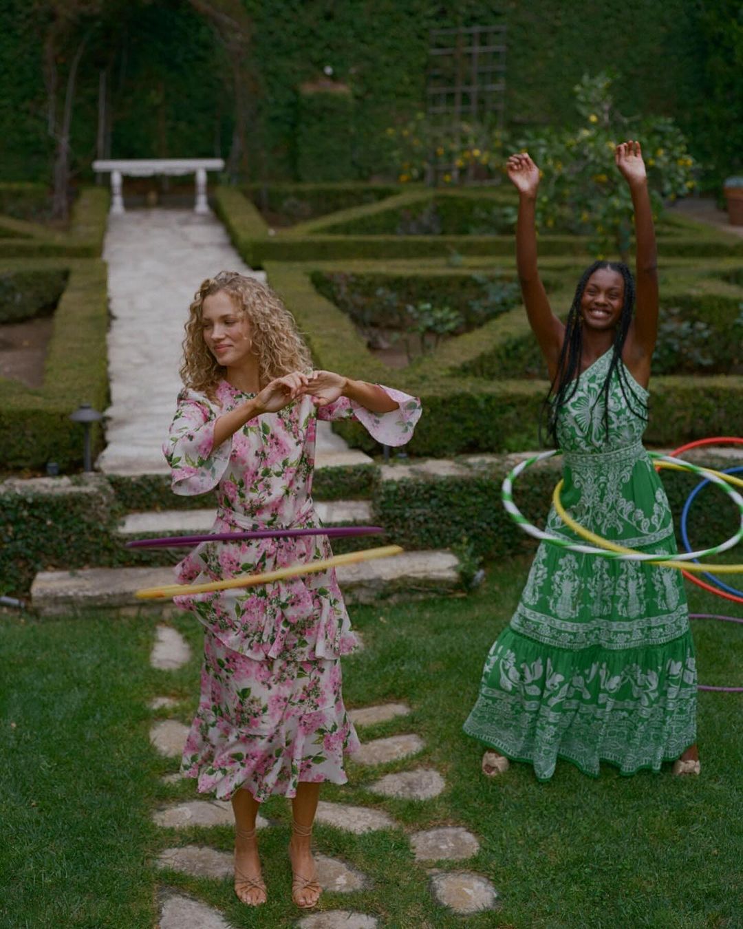Two women in floral dresses play with hula hoops on a lawn in a garden setting with trimmed hedges and a stone pathway.