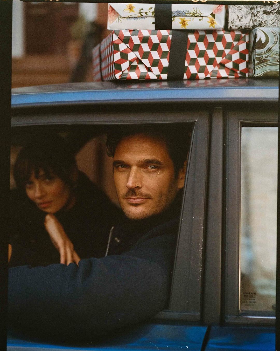 Handsome man in a blue car with a woman, packages on the roof, medium shot, portrait orientation, outdoor setting, lifestyle, fashion editorial.