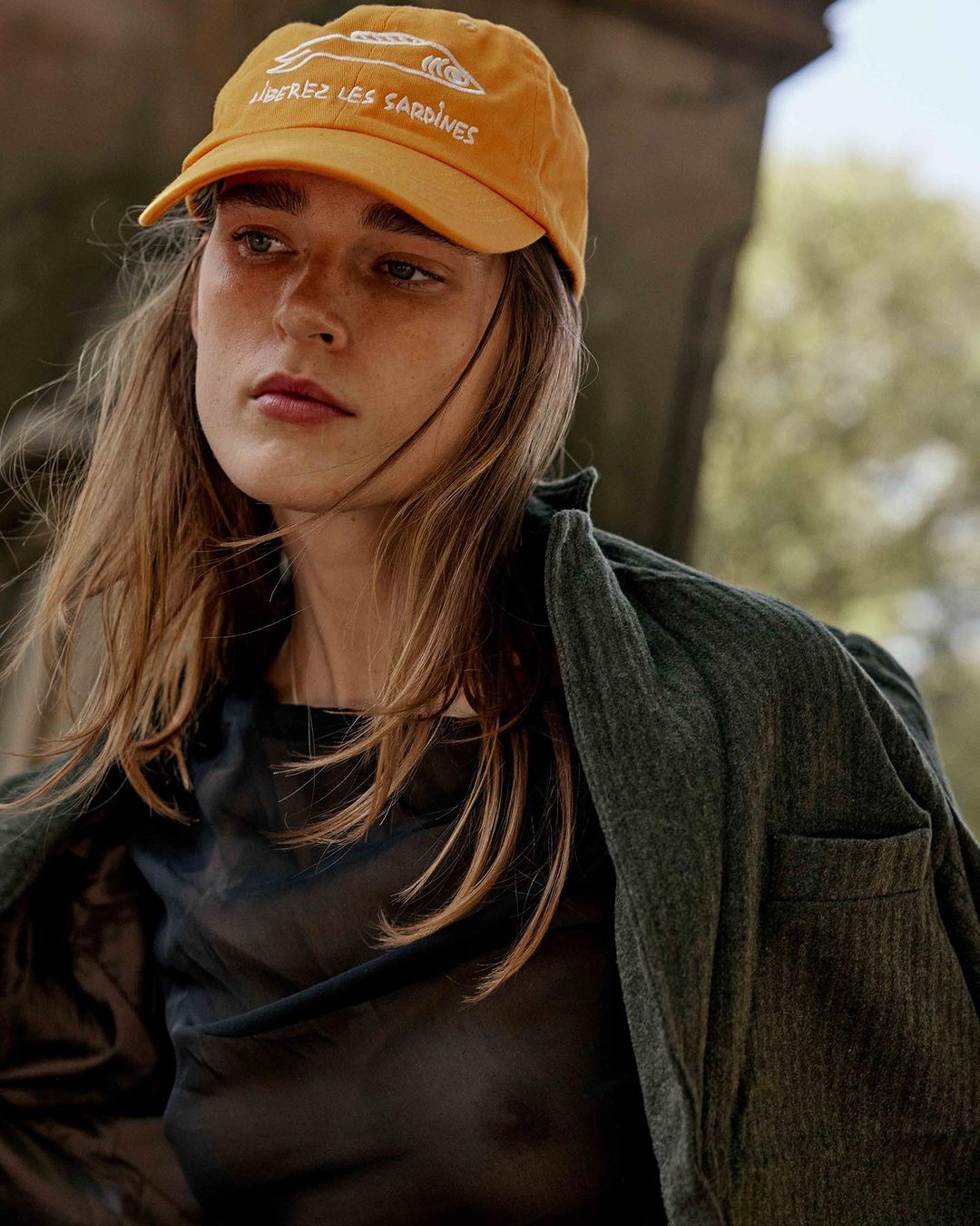 Woman wearing a yellow baseball cap with a graphic and text, and a green jacket stands outdoors in a portrait shot.