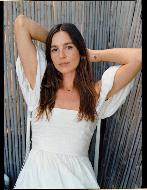 Adult woman with arms raised, wearing a white dress sitting on a chair in front of a bamboo fence in a medium portrait shot.
