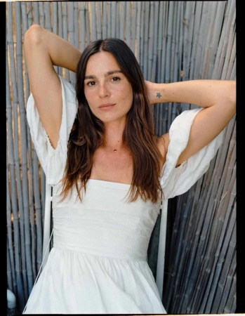 Adult woman with arms raised, wearing a white dress sitting on a chair in front of a bamboo fence in a medium portrait shot.